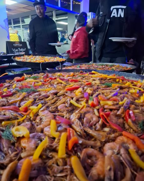 Large seafood paella pans being served fresh at a corporate function, topped with prawns, peppers, and herbs while guests gather around the live cooking station.