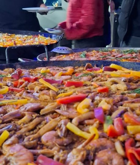 Large seafood paella pans being served fresh at a corporate function, topped with prawns, peppers, and herbs while guests gather around the live cooking station.